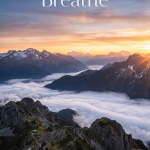 Mountain sunrise wallpaper with rocky foreground, clouds in the valley, warm sky colors, and the text ‘Breathe’